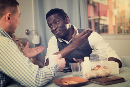 Cheerful African American man talking and laughing with friend while sitting at home tableの写真素材