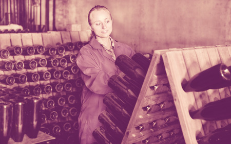 Portrait of smiling female wine maker wearing coat taking care of seasoning bottles at winery cellarの写真素材