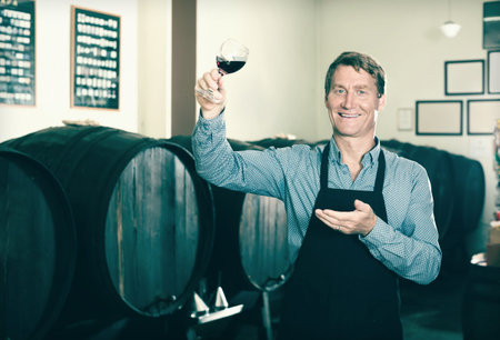 man seller wearing uniform holding glass of wine in wine cellar