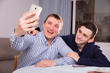 Guy with his father are resting and taking selfie by phone at the home.