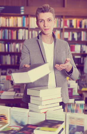 Young cheerful positive salesman of bookshop holding fat book in hand, offering it to somebodyの写真素材