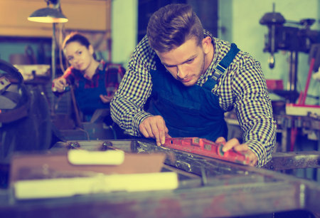 concentrated young master is standing near workplace and working with level in workshop.