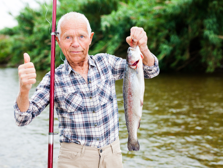 Senior caucasian fisherman satisfied with fishing holding large trout in handの写真素材