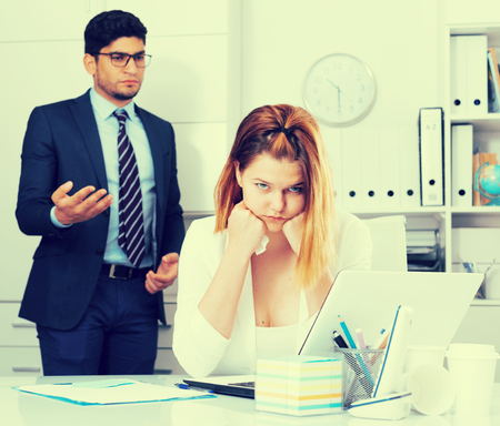 Frustrated business woman sitting at office desk on background with angry chiefの写真素材