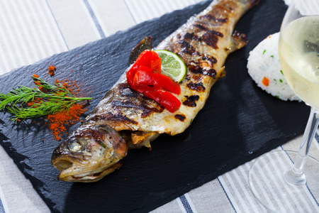 Delicious baked trout fish served with white wine on black board with garnish of white rice, grilled bell pepper, dill and lemon sliceの写真素材