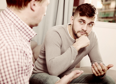 Anxious man discussing problems with friend while sitting on sofa at homeの写真素材