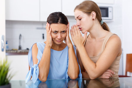 Portrait of sad young  girlfriends whispers in the ear and sharing secrets at table