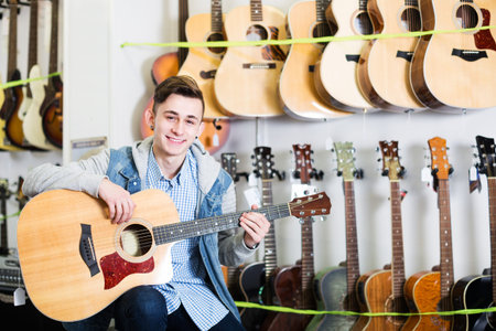 Positive male teenager examining various acoustic guitars in guitar shop