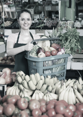 Smiling attractive saleswoman with shopping basket with greengrocery at farmers marketの写真素材