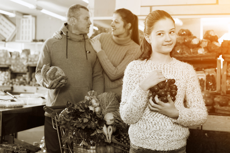 Cheerful tweenager girl holding big cluster of red grapes during family shopping in fruit storeの写真素材