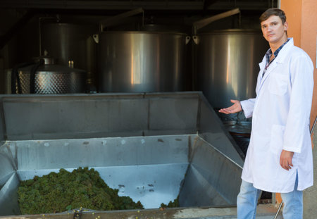 Young male owner of winery standing near grapes crusher machine, showing process of winemaking