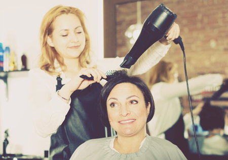 Happy young hairdresser styling hair to adult woman with hairdryer and brush in hairdress salonの写真素材
