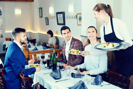 Polite cheerful positive smiling waitress bringing ordered dishes to guests at restaurantの写真素材