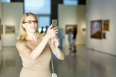 Woman wearing glasses visiting picture gallery and taking photo on her smartphoneの写真素材