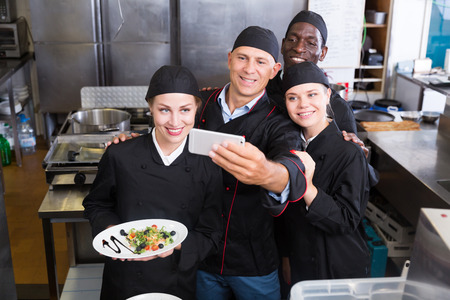 Cheerful friendly group of chefs taking selfie in modern kitchen of restaurantの写真素材