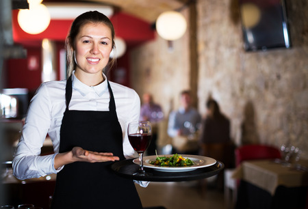 Smiling waitress wearing apron holding tray with dishes in restaurant interiorの写真素材