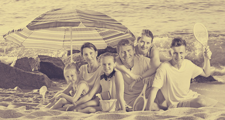 Cheerful parents with four children in different ages and beach toys sitting under the sun umbrellaの写真素材