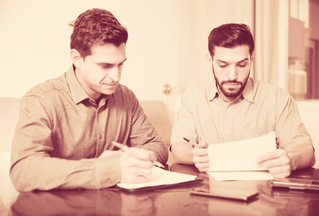Two serious male friends reading attentively and discussing documents while sitting at home tableの写真素材
