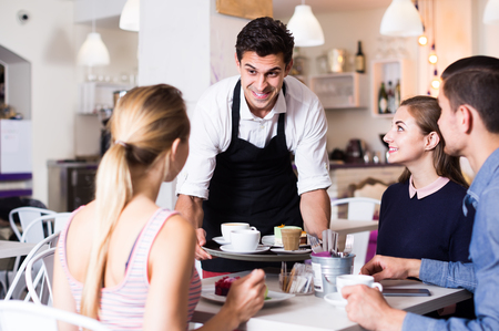 Polite happy waiter bringing ordered dishes to friends in tearoom of a cozy confectioner's shopの写真素材
