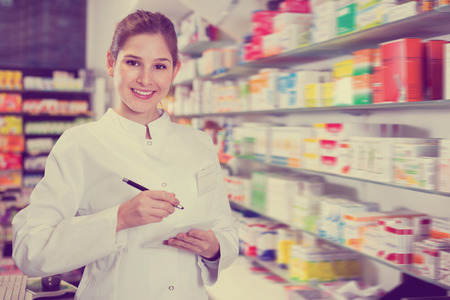 Diligent positive female specialist is attentively stocktaking medicines with notebook near shelves in pharmacy.の写真素材