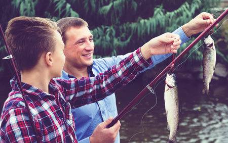 Portrait of happy father with teenager son looking at fish on hook in hands outdoors