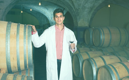 professional taster of winery in white robe standing with glass of red wine in wine cellar
