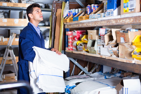 Diligent friendly  sellerman in uniform is putting bags on the shelves in the building store.の写真素材