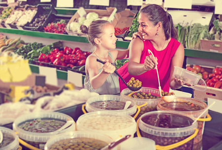 Smiling woman with girl taking pickled olives from bucket in food shopの写真素材
