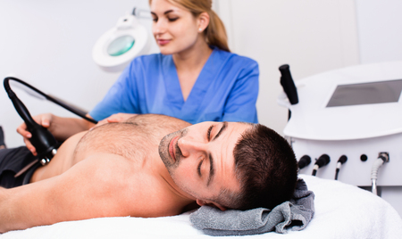 Qualified glad cheerful woman cosmetologist with male client during hardware body procedure in modern aesthetic clinicの写真素材