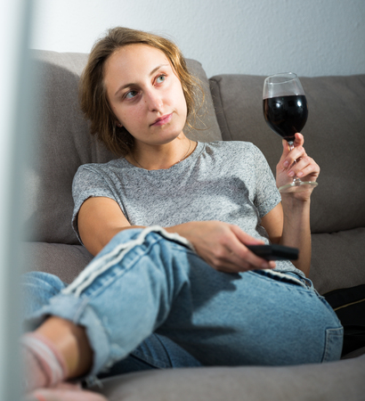 Smiling young woman with glass of wine and TV remote sitting on couch at home watching movieの写真素材