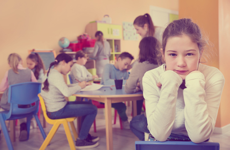 Upset girl standing at a schoolroom on background with pupils studying with teacherの写真素材