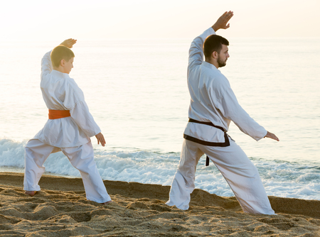 Man and boy in uniform practising karate poses at sunny sea beachの写真素材