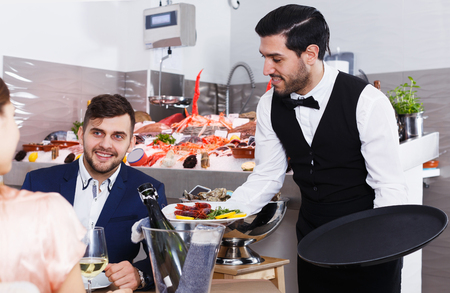 Polite attractive waiter bringing ordered seafood dishes to smiling couple at fish restaurantの写真素材