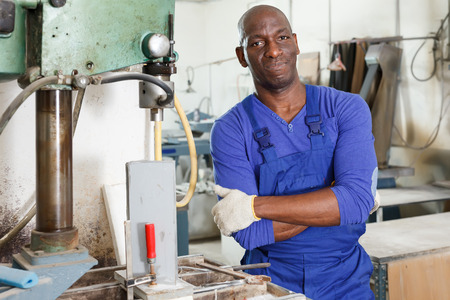 Confident African American glazier working with glass on stationary drilling machine in workroomの写真素材