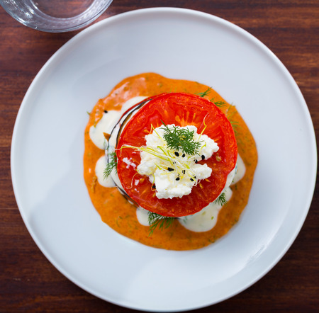 Top view of fried eggplant pyramid with tomatoes on spicy sauce with soft cheese and greens served on white plate on wooden backgroundの写真素材