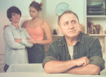 Woman and her daughter are sympathying their sad father who is sitting at the table at home.の写真素材