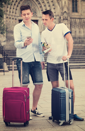 Happy positive smiling male couple  with luggage using the map at smartphoneの写真素材