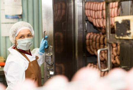 Woman checking quality of cooked smoked sausages in professional baking machine at food production factoryの写真素材