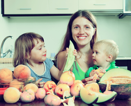 Happy woman with daughters eating melon and peaches at homeの写真素材