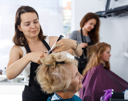 Skilled woman hairdresser making hairstyle for elderly female client in hair studioの写真素材