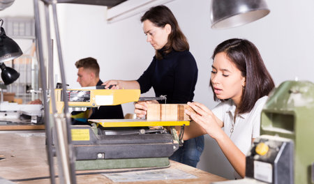Focused Brazilian girl working in architecture modeling workshop, using scroll saw for making...