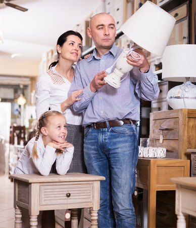 Positive family couple with their daughter are choosing lamp in furniture storeの写真素材
