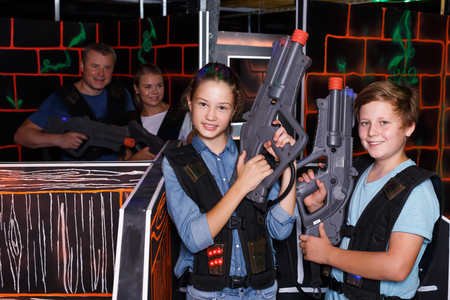 Ordinary teenage boy and girl posing with laser guns while having fun with adults on laser tag arenaの写真素材