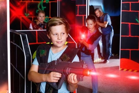 Excited glad cheerful  smiling boy aiming laser gun at other players during laser tag game in dark roomの写真素材