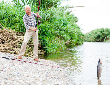 Portrait of positive senior man holding fishing tackle and catch fish on hook at riverside