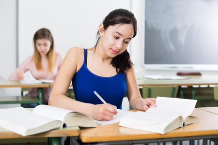 Girl is sitting at the desk and writing homework in the classroom.の写真素材