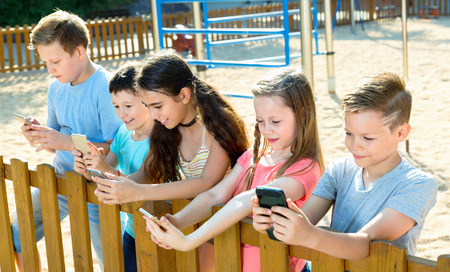 Five glad  cheerful smiling children sitting and playing in smartphone at the playgroundの写真素材