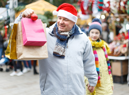 Happy man wearing warm clothes and Santa hat holding bags after shopping with daughter on outdoor Christmas marketの写真素材