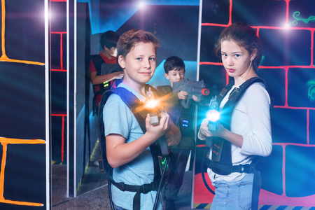 Cheerful  smiling teen girl and boy with laser pistols playing laser tag with friends on dark labyrinthの写真素材