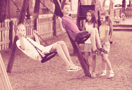 Positive girls and boys in school age swinging on children's playground outdoorsの写真素材